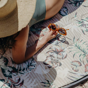 Basil Bangs beach rug in Tiki print with beige and white stripes and tropical leaf print. A woman holding sunglasses, in a wide brim straw hat is lying back on her elbows in the sun. 