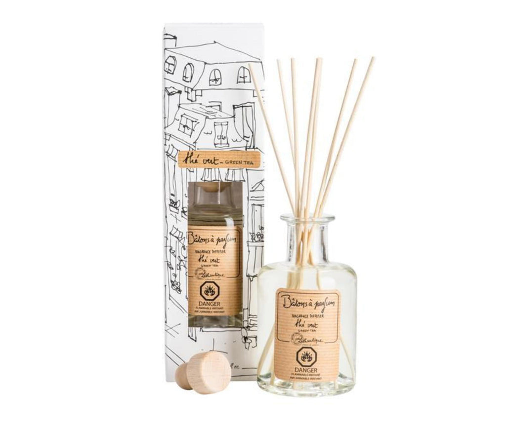 A clear, glass bottle of Green Tea scented diffuser, with 7 bamboo reeds in the neck of the bottle and a lable printed with " Batons a parfum, fragrance diffuser, the vert, green tea" and the Lothantique logo. Shown with a second bottle in white packaging with black line drawings of French houses.