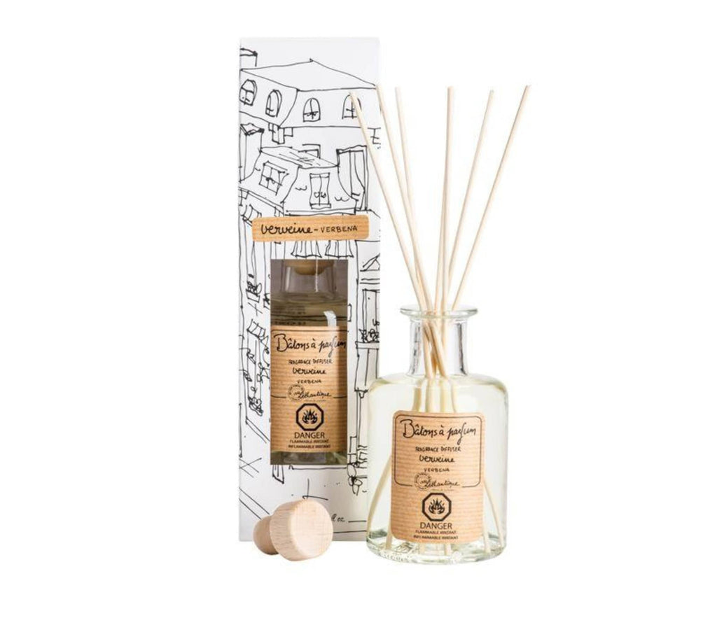 A clear, glass bottle of verbena scented diffuser, with 7 bamboo reeds in the neck of the bottle and a lable printed with "Batons a parfum, fragrance diffuser, verveine, verbena" and the Lothantique logo. Shown with a second bottle in white packaging with black line drawings of French houses.