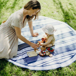 Basil Bands round beach blanket, in Atlantic with blue and white stripes, shown on grass with a small child and a woman having a snack in the sun.