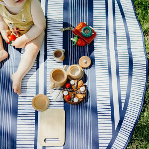 Basil Bands round beach blanket, in Atlantic with blue and white stripes, shown on grass in a close up with a small child having a snack in the sun.