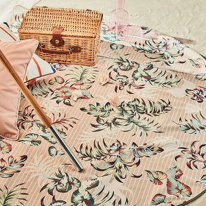 Basil Bangs beach blanket in Tiki print with beige and white stripes and tropical leaf print, spread out on the sand with a wicker picnic basket and a two outdoor cushions.
