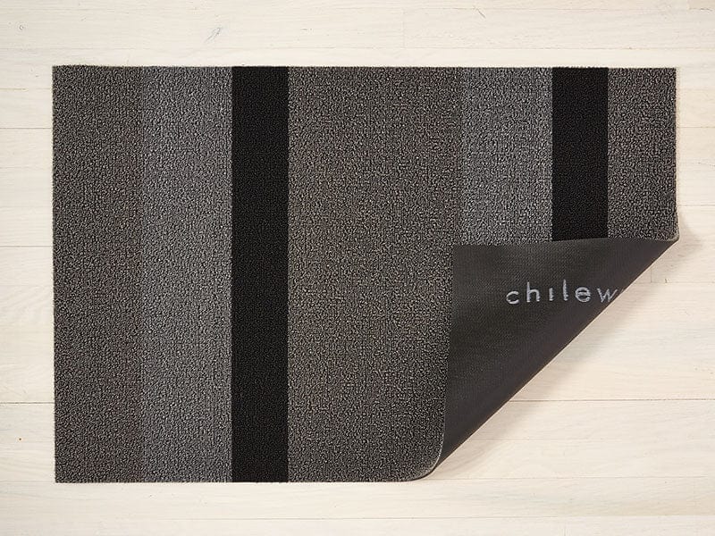 a rectangular, bold striped shag floor mat in black and shades of grey made of eco friendly looped vinyl yarns backed with commercial black rubber