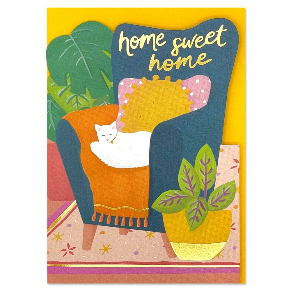 an illustrated greeting card featuring a colourful living room scene with a white cat asleep on a blue armchair, text in gold foil reads "home sweet home"